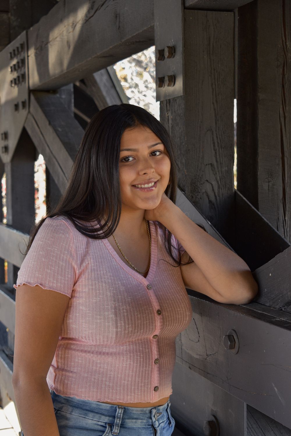 Smiling young woman in pink top leaning against wooden structure outdoors.