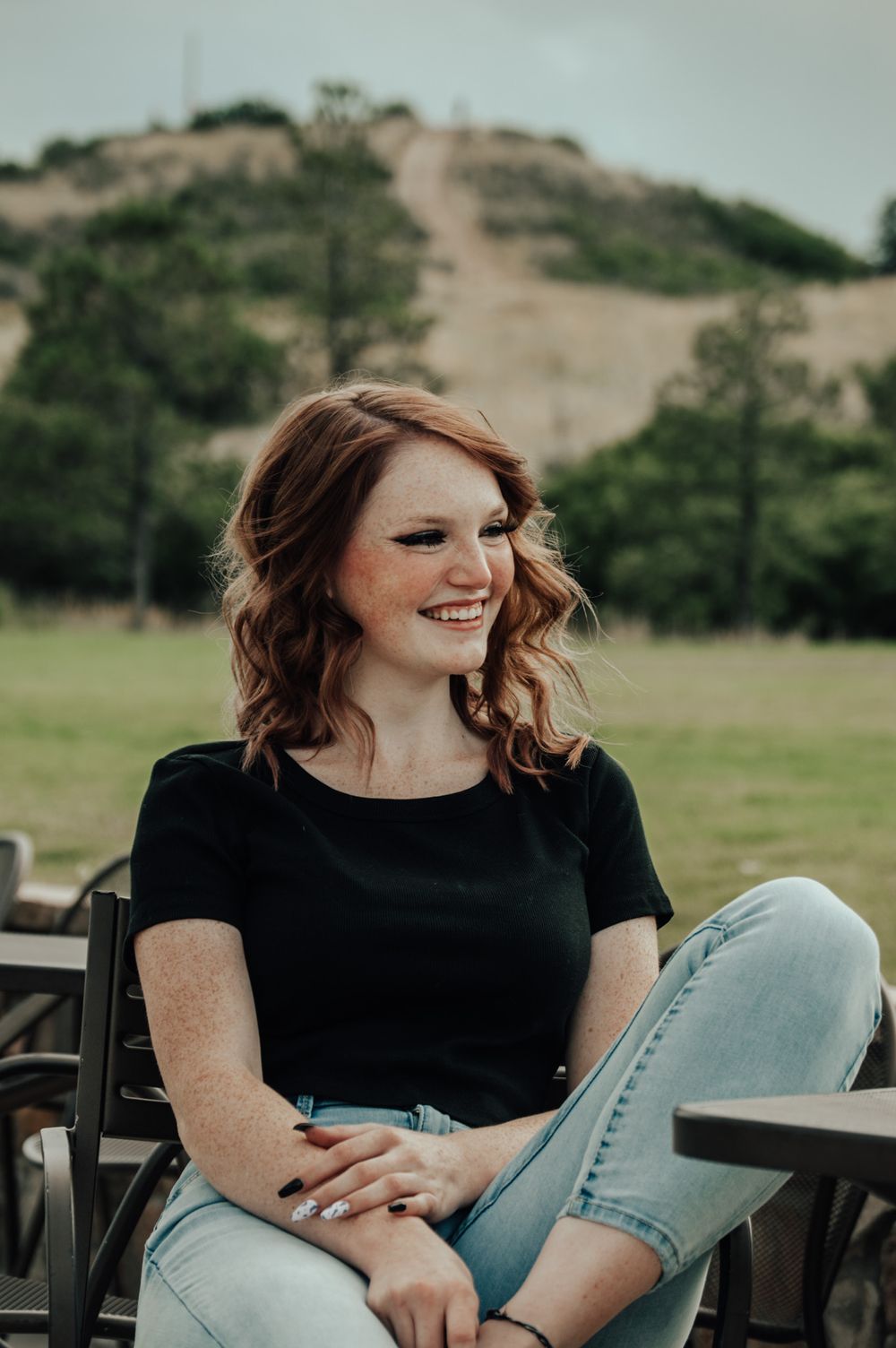 Smiling redhead woman in black t-shirt and jeans seated outdoors with green hills behind.