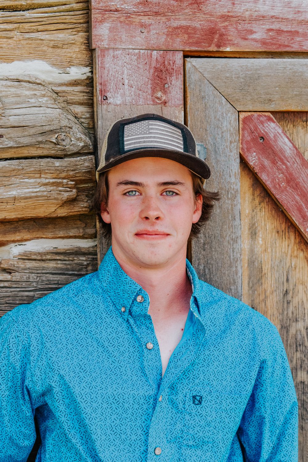 Young man in blue patterned shirt and cap leaning against a rustic wooden barn.