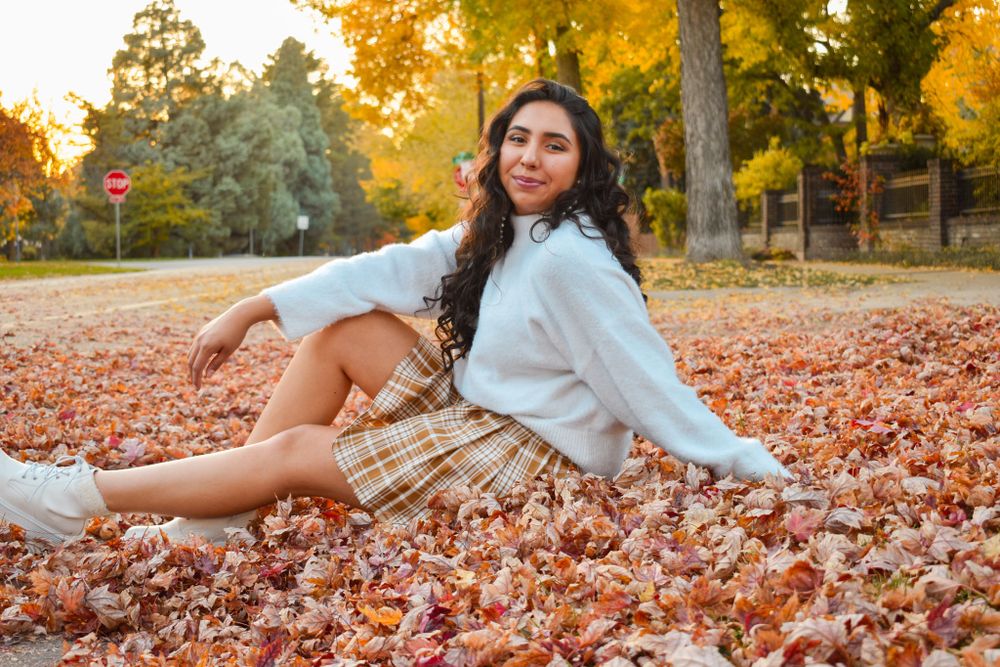 A young woman in a white sweater and plaid skirt sits in colorful autumn leaves outdoors.
