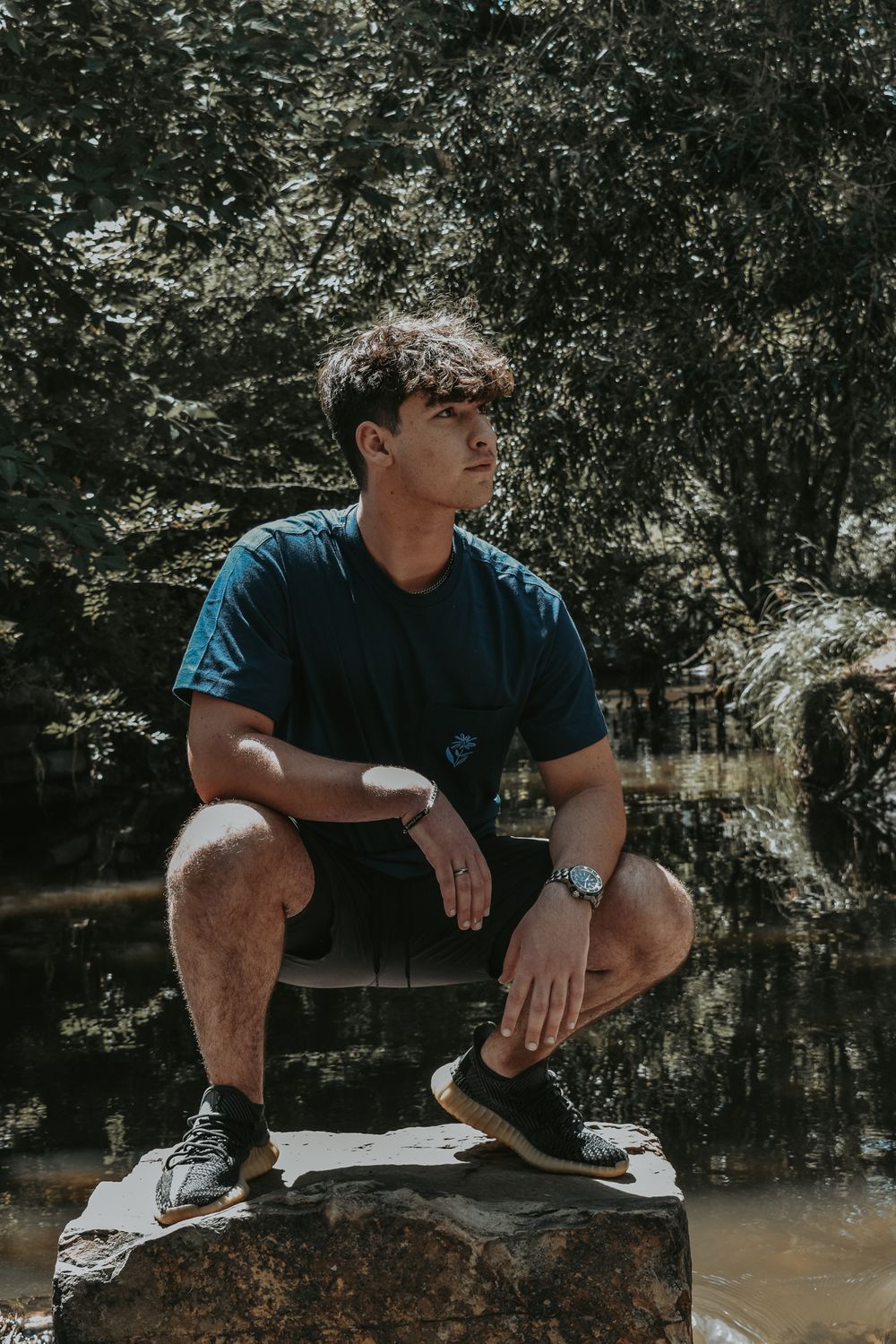 Young man in blue shirt and shorts crouching on a rock near a pond.