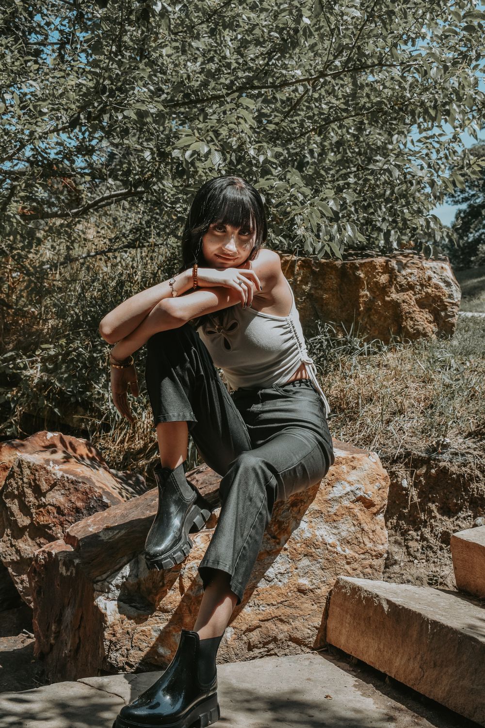 Young woman in white tank top and dark jeans posing on rocks outdoors.