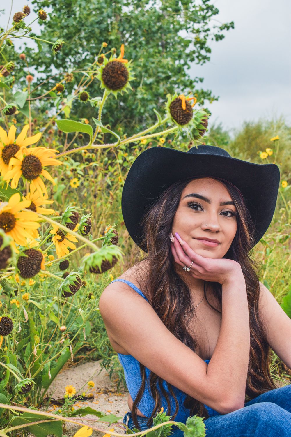 Young woman in black cowboy hat sitting among sunflowers in a field.