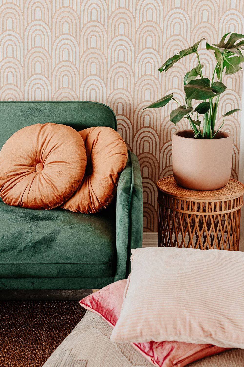 Cozy living room corner with emerald green velvet couch, peach round pillows, and rattan side table against patterned wallpaper.