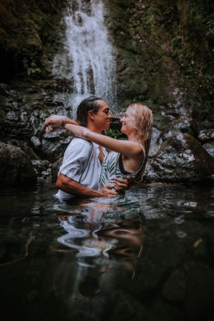 A couple playing in the water beneath a cascading waterfall.