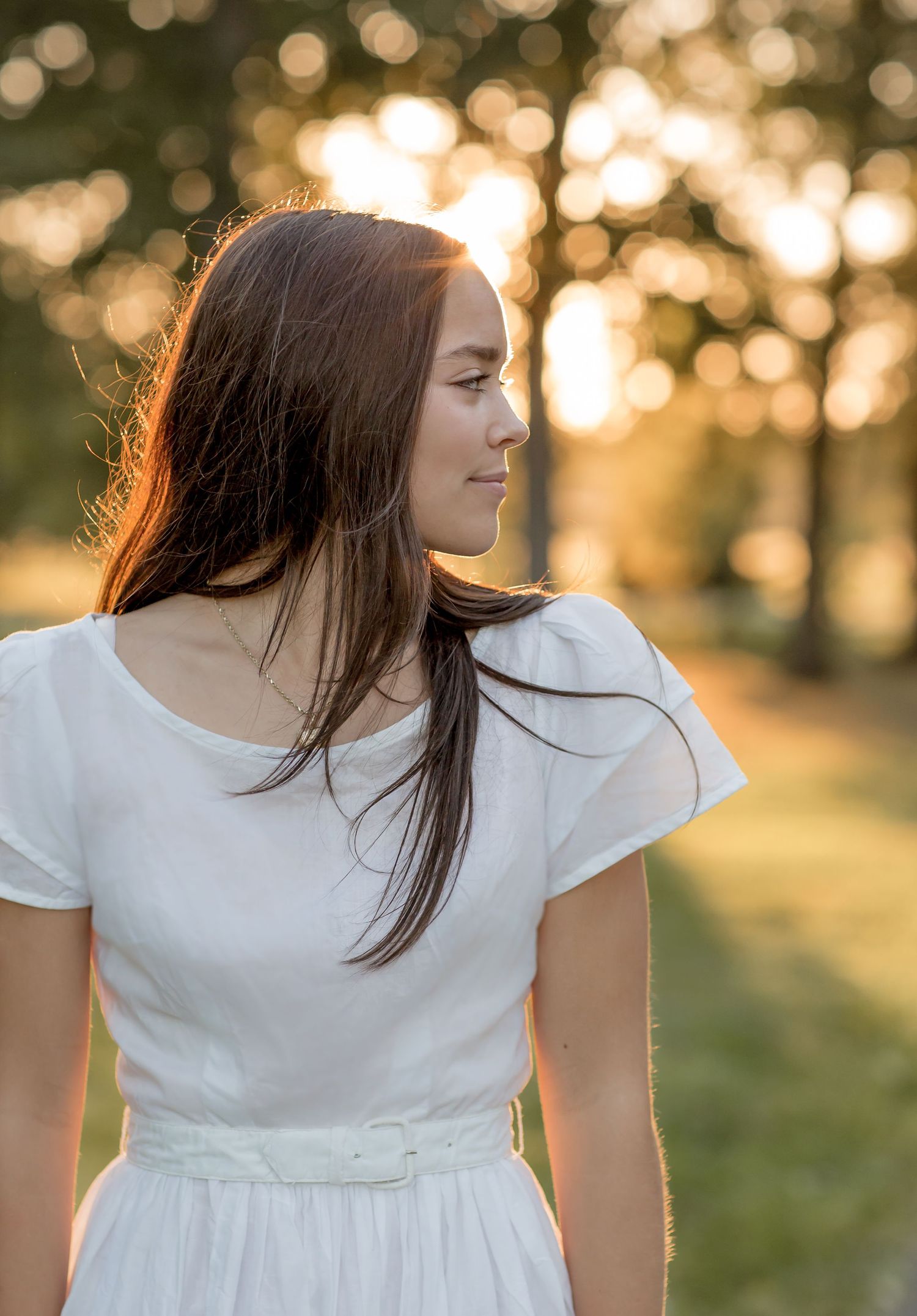 Young brunette woman in white belted dress gazes sideways in a glowing golden hour outdoor portrait.