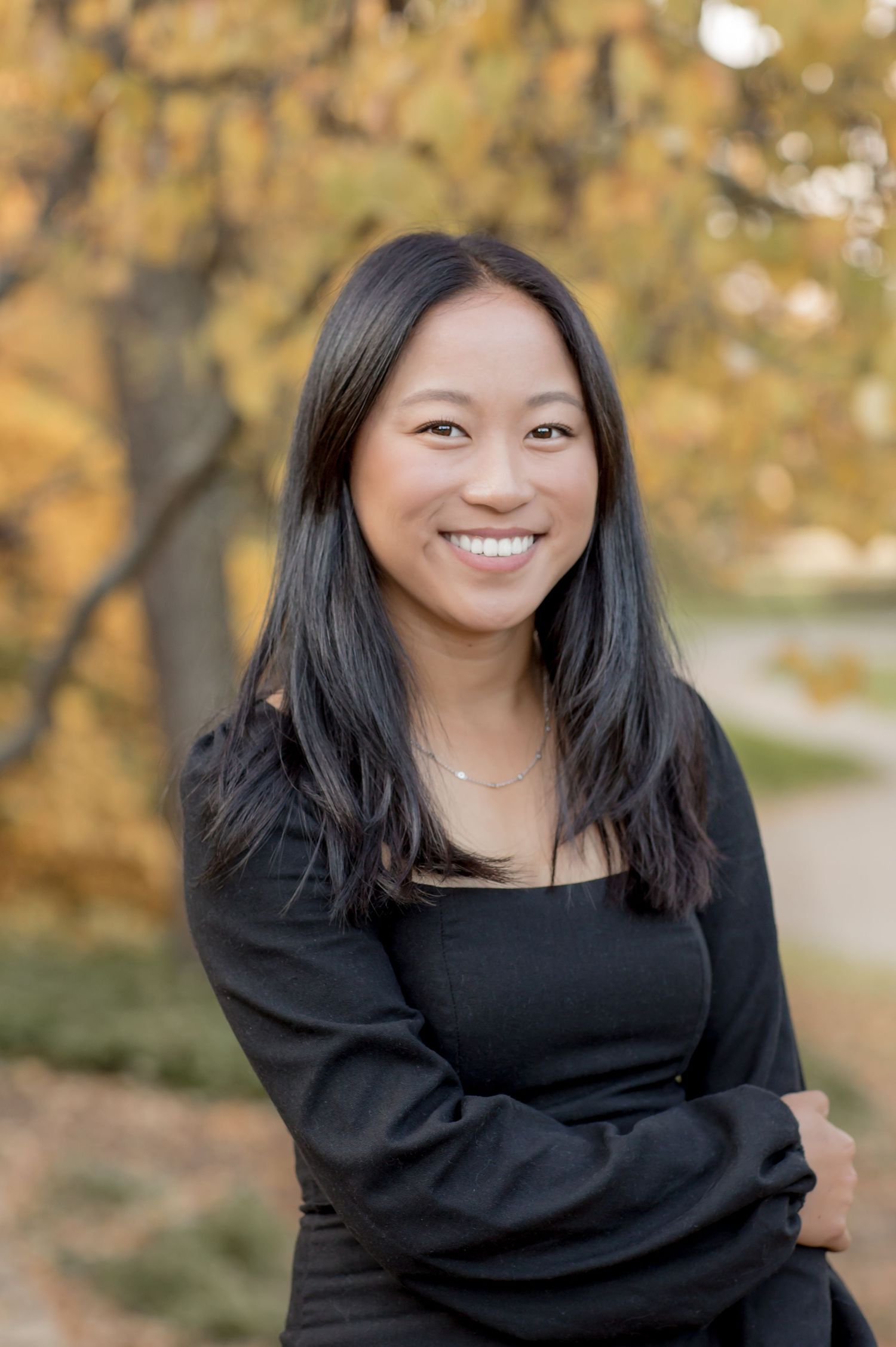 Smiling young Asian woman in black outfit posing outdoors with autumn foliage in the background.