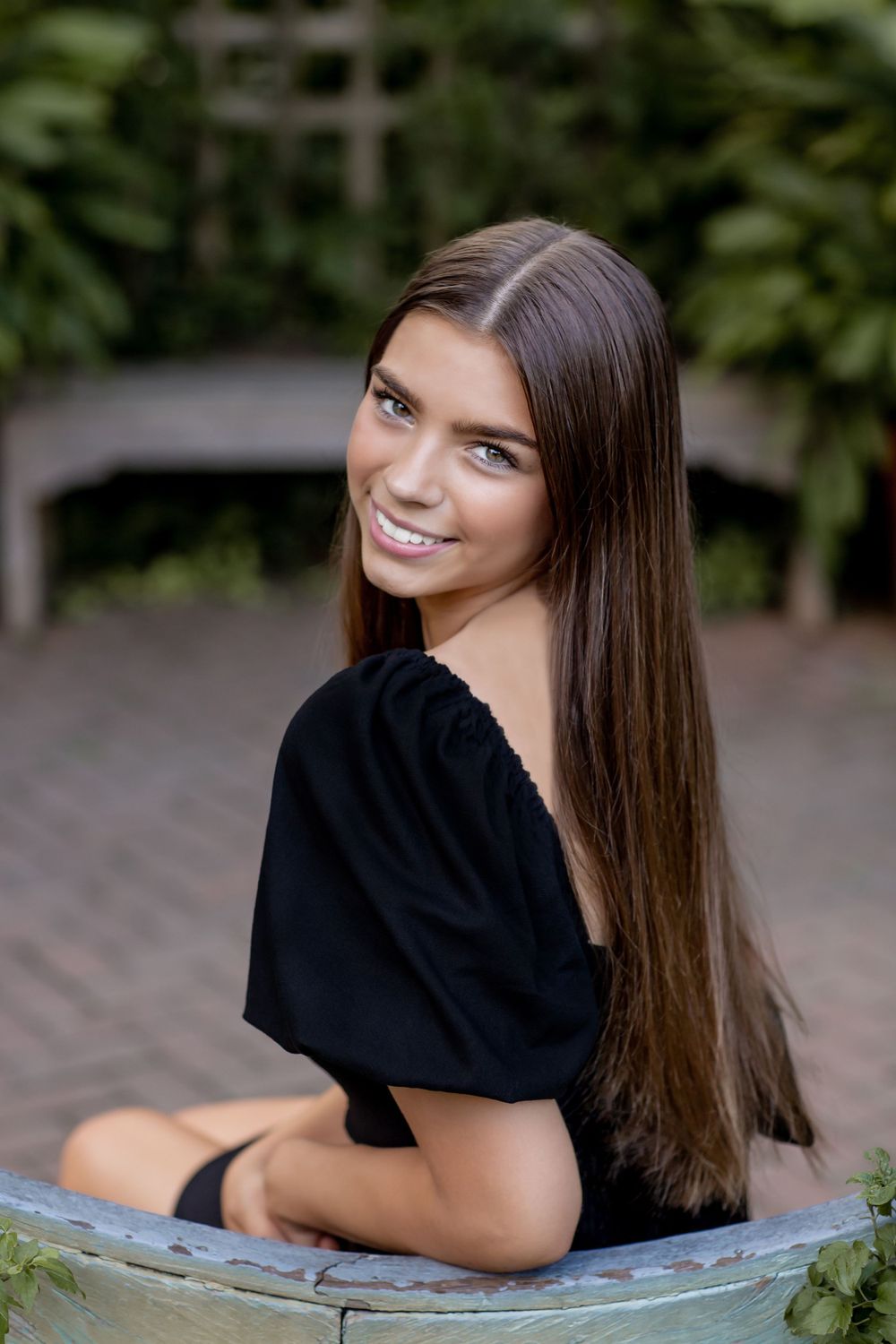 Young woman with long brown hair in a black top smiling over her shoulder in a garden setting.