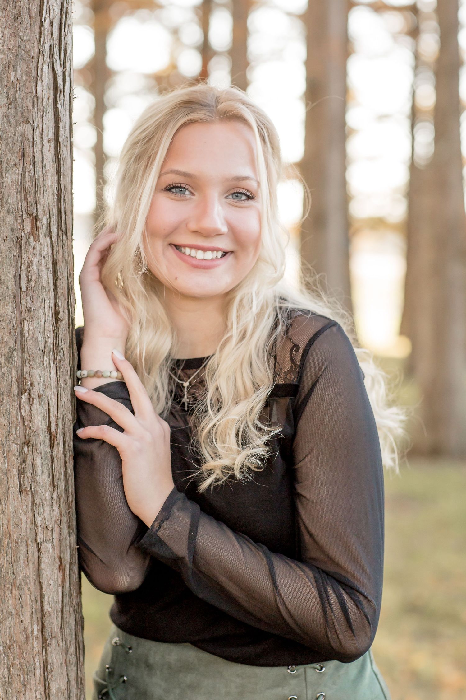 Smiling blonde teen girl in black sheer top leans against a tree in a pine forest senior portrait.