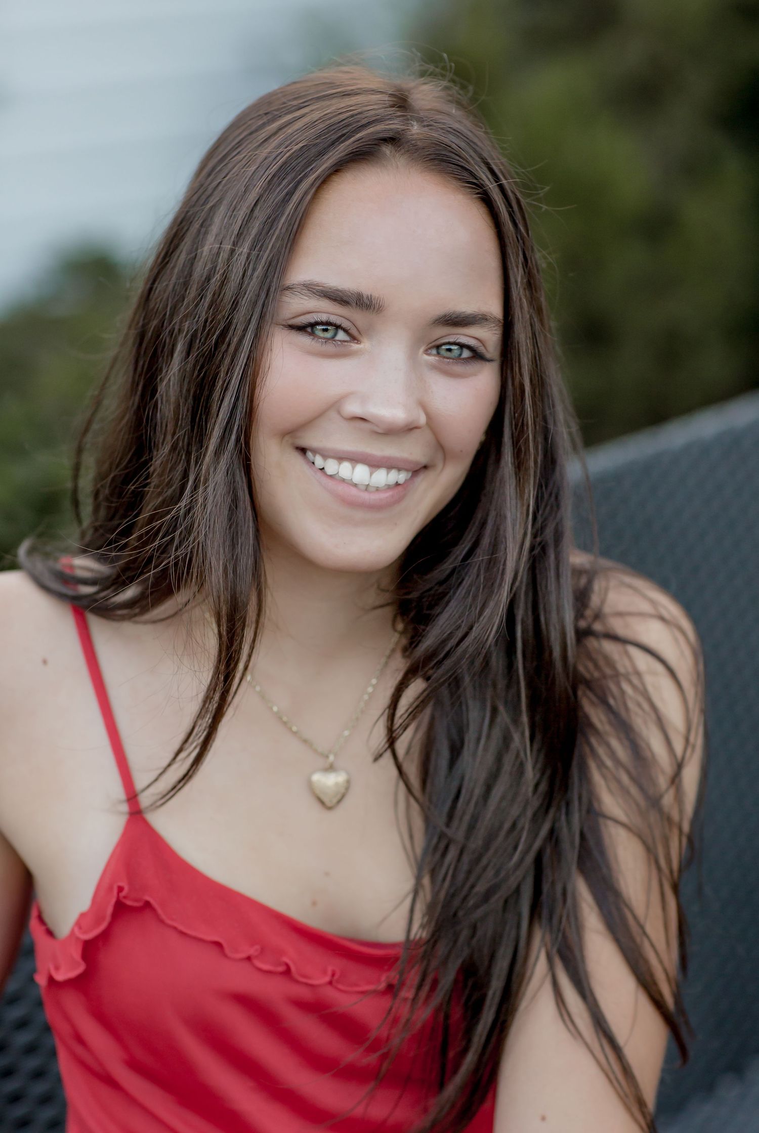 Smiling young woman with long brown hair wearing a red dress and gold heart necklace outdoors.