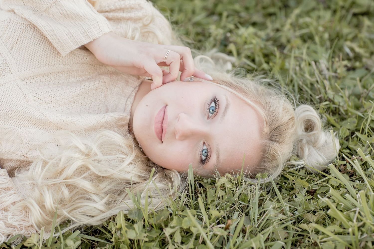 Blonde girl with striking blue eyes lies in green grass looking up in a dreamy outdoor portrait.