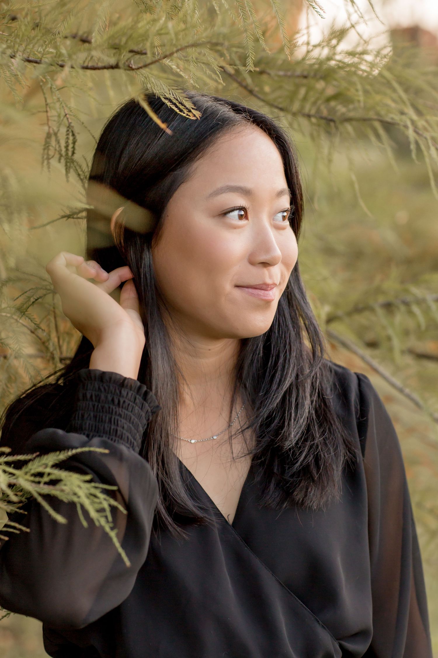 Young Asian woman in black blouse standing near pine branches looking away thoughtfully.