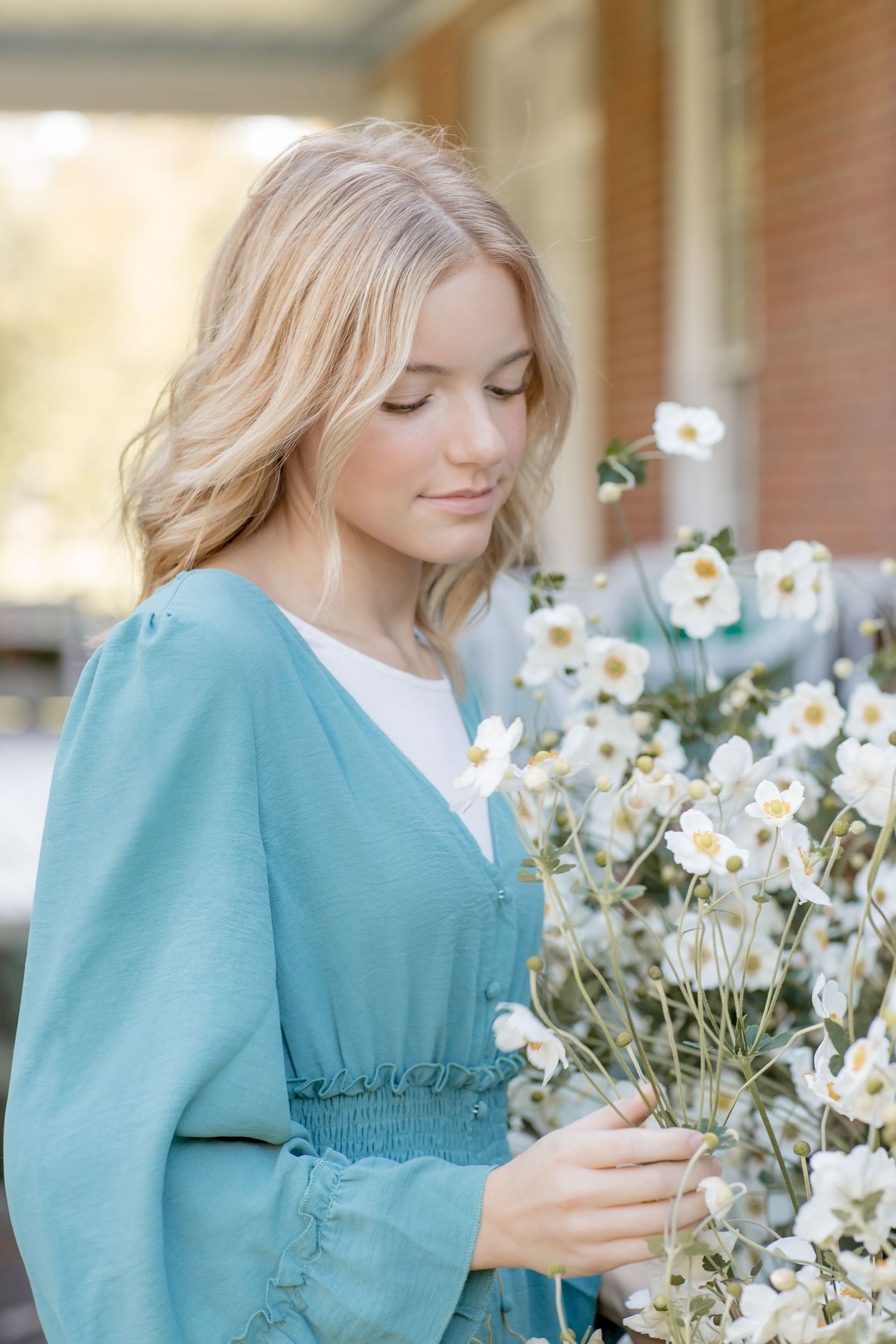 Blonde young woman in teal dress gently touching white flowers in a soft natural light setting.