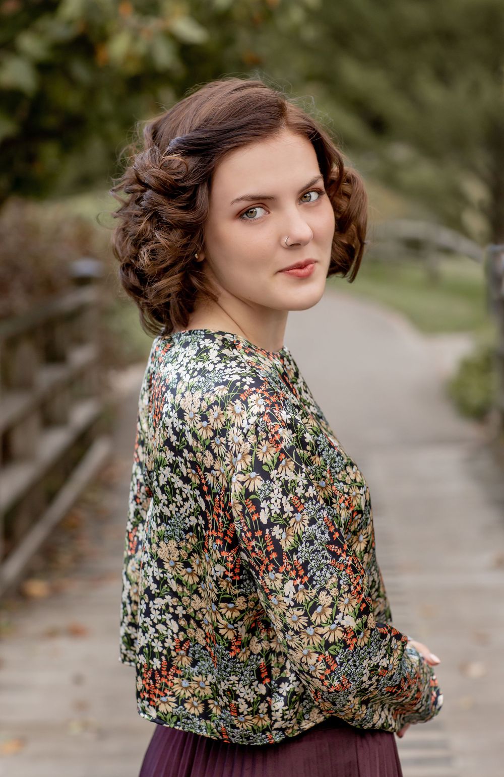Young woman with elegant updo hairstyle wearing a floral blouse, posing on a rustic bridge in autumn.