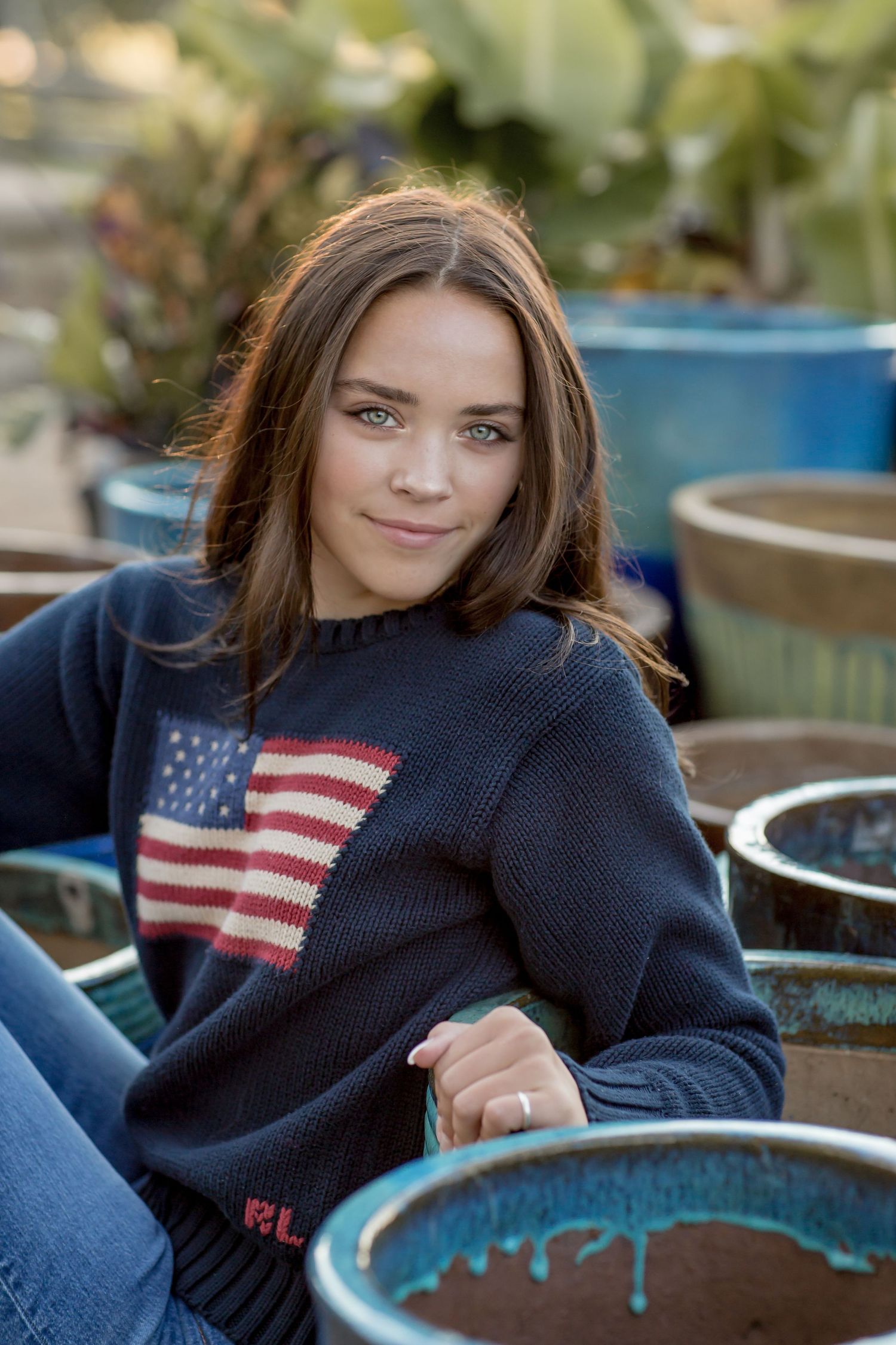 Young woman in Ralph Lauren American flag sweater sitting among terracotta pots in a garden.