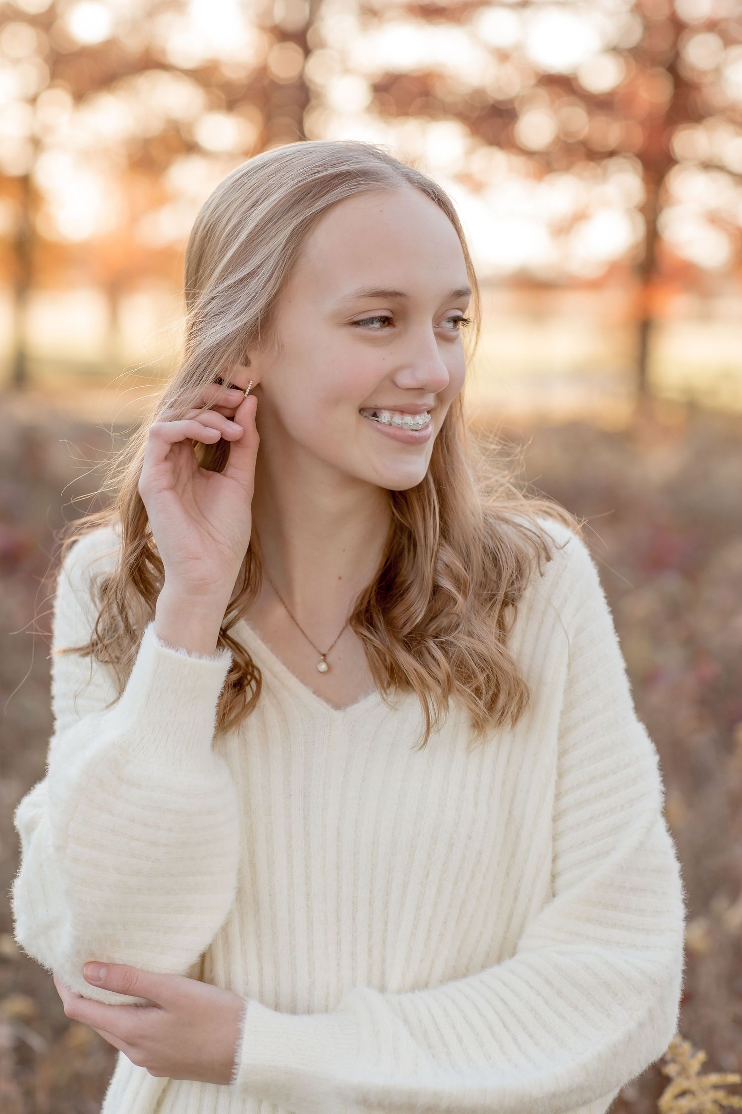 Smiling blonde young woman in cream sweater touches her hair in warm autumn outdoor setting.
