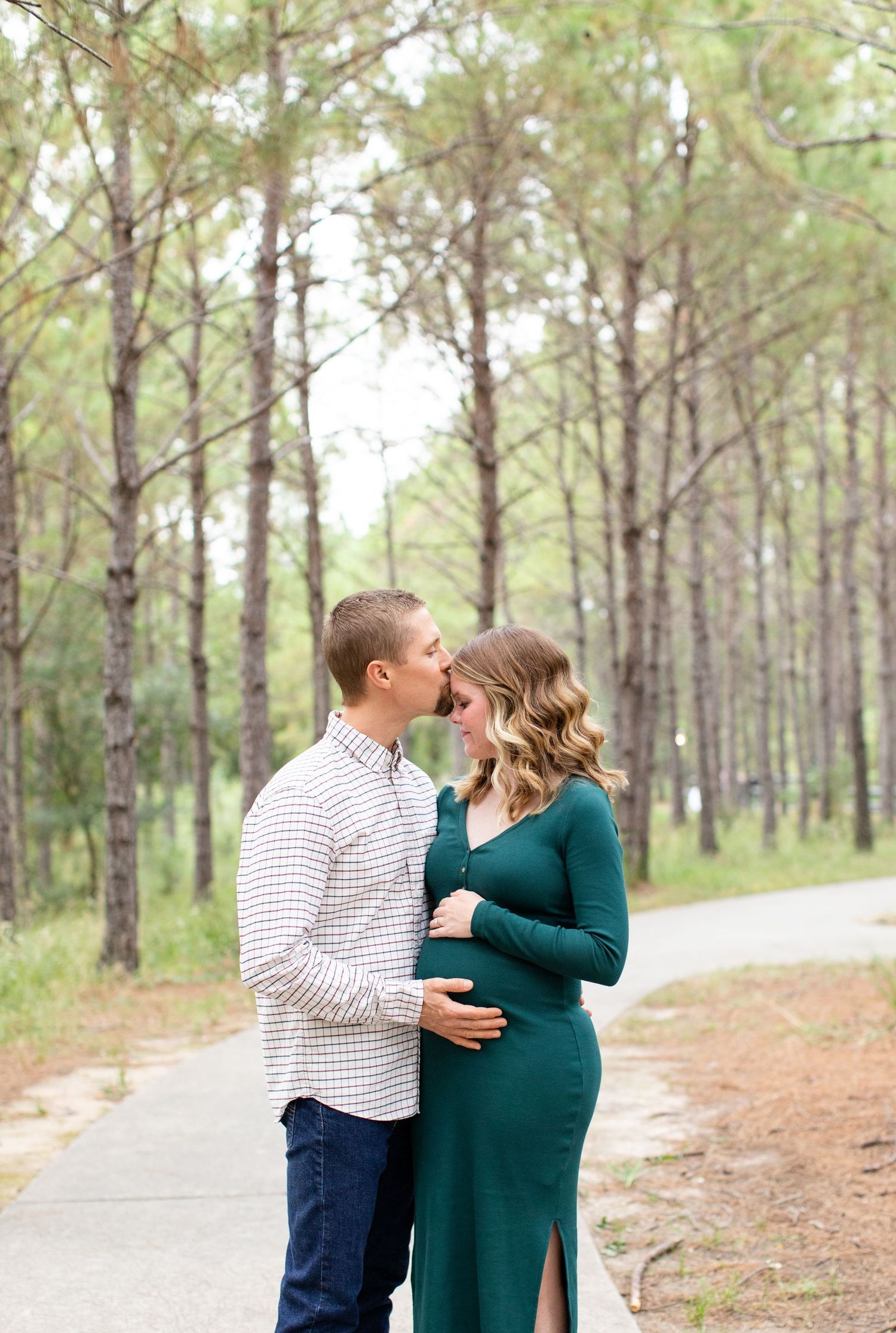 Romantic pine forest maternity photo with couple embracing on a winding path.
