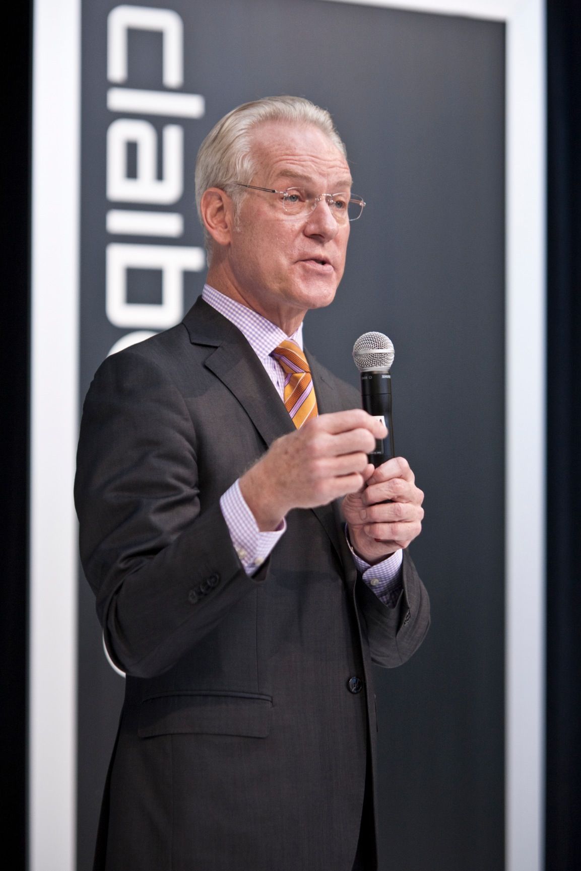 Business speaker in suit and tie presenting with microphone against dark backdrop.