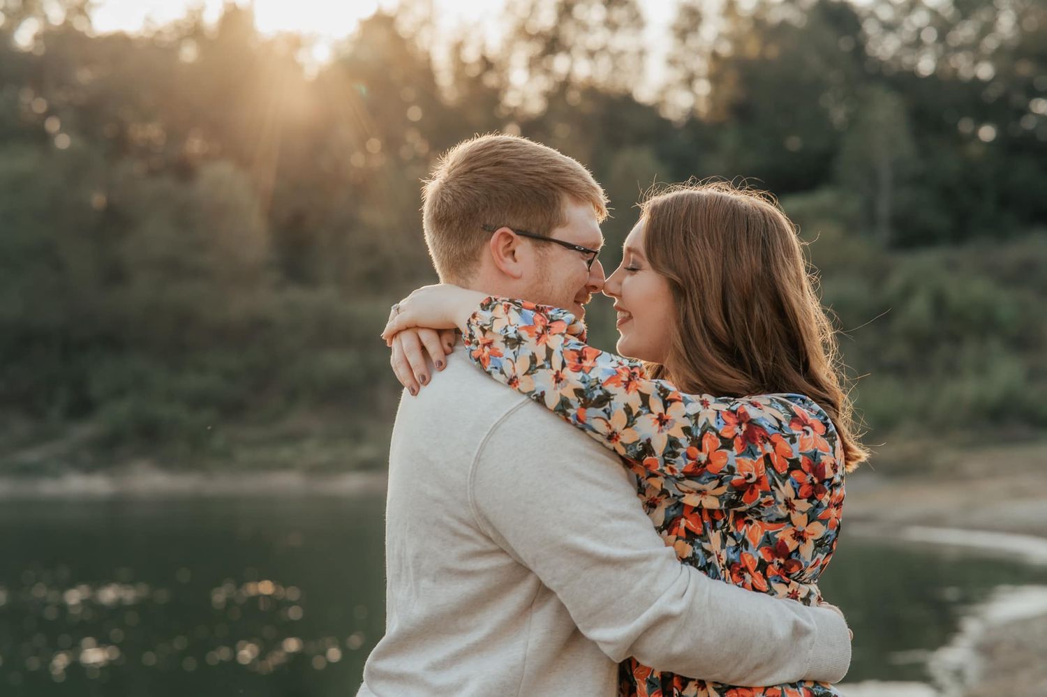 A romantic couple embraces at sunset near a lake with golden sunlight filtering through trees in the background.