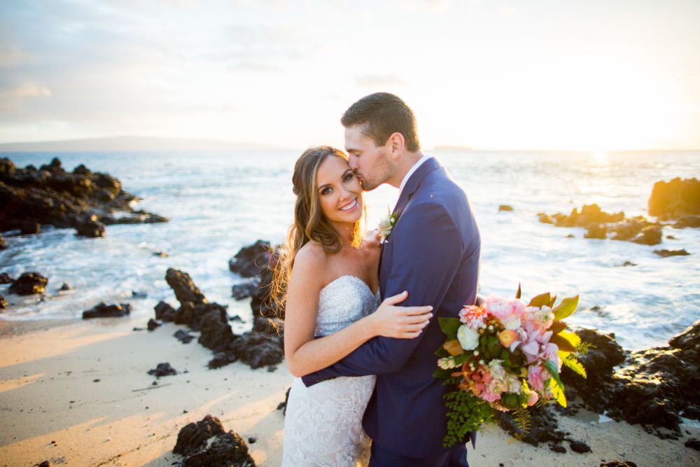 A romantic sunset beach wedding photo with crashing waves and rocky coastline in the background.