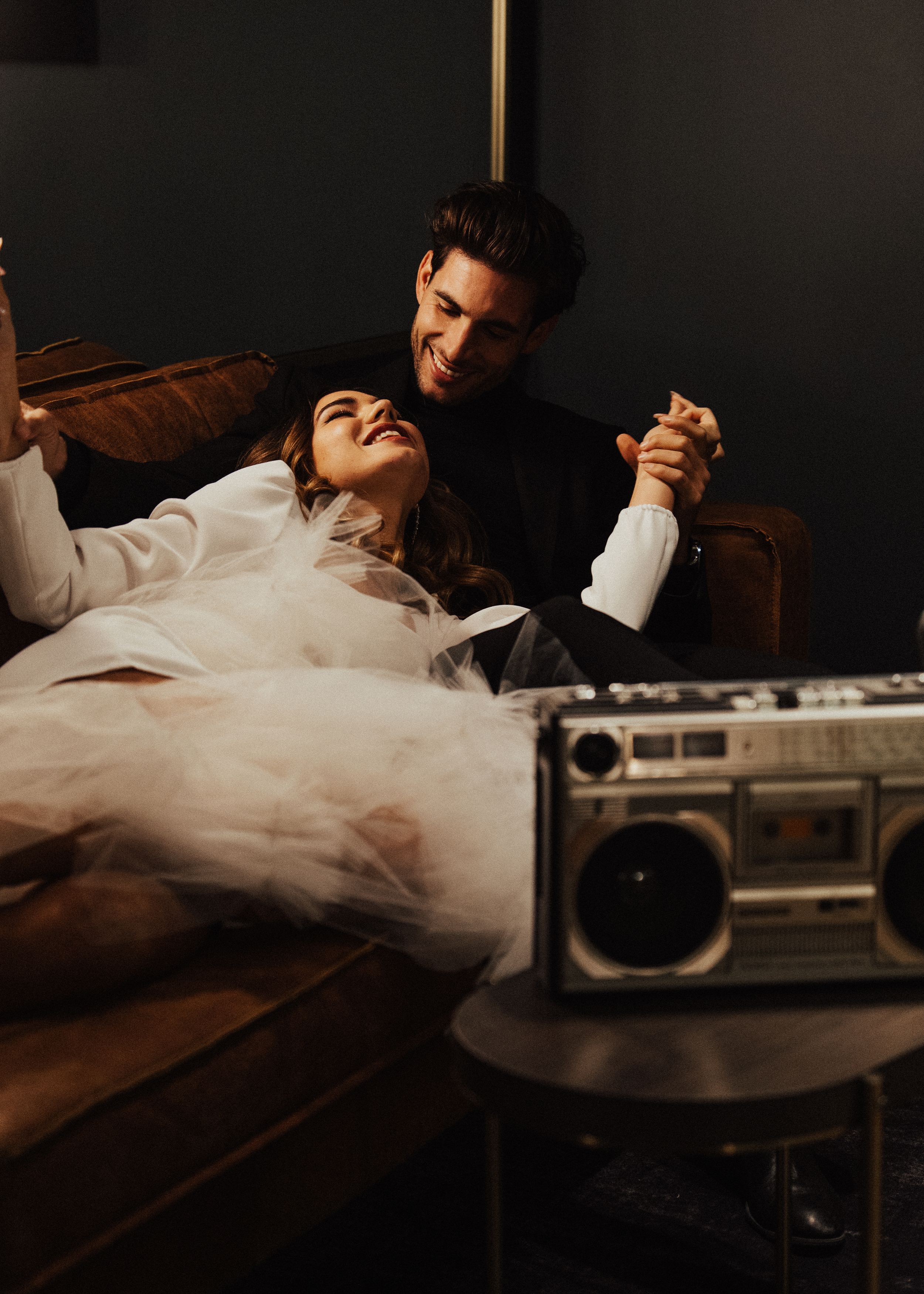 Artistic wedding photos with couple lounging near vintage boombox in dark setting.