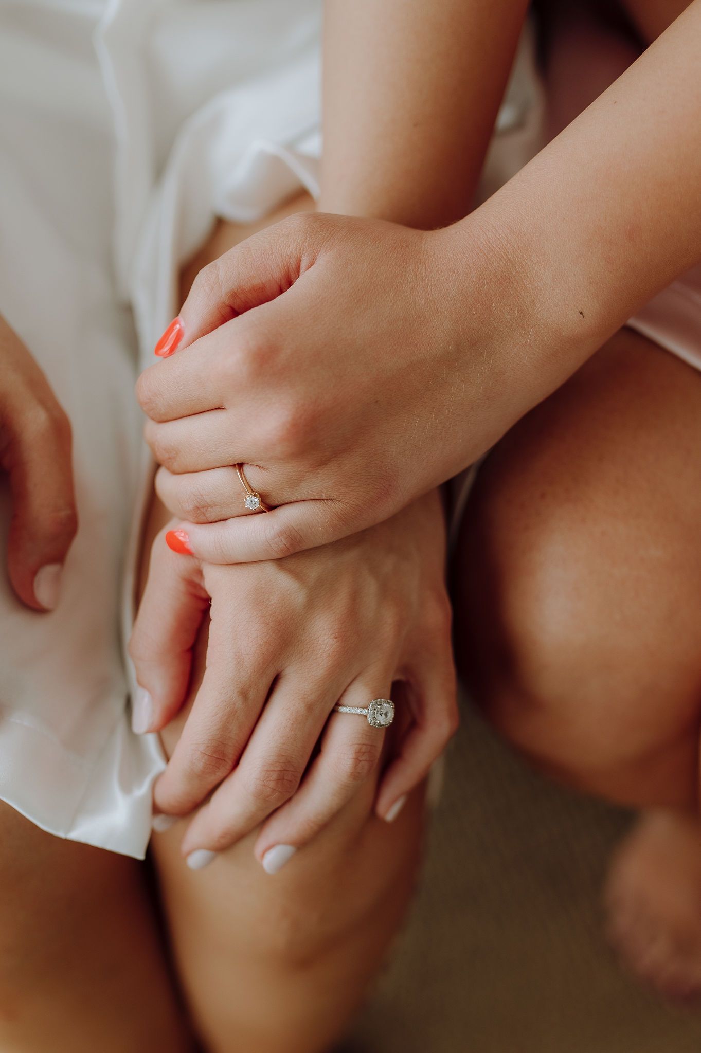 Close-up of hands with engagement ring and coral nail polish against white fabric.