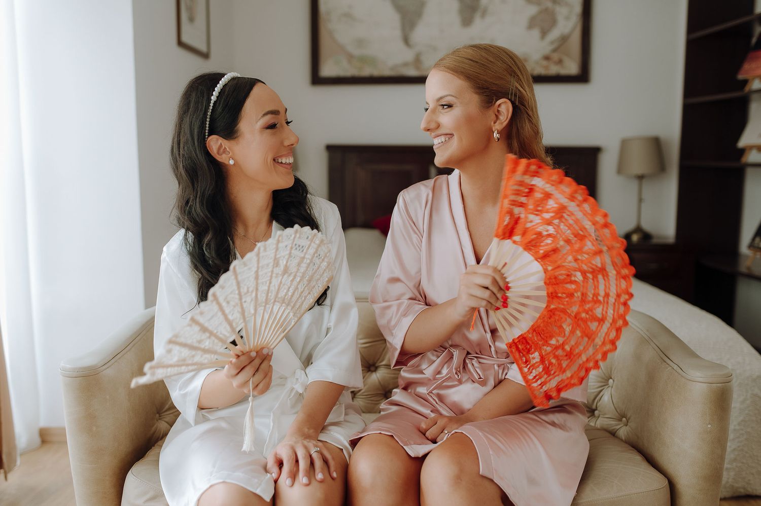 Two friends sharing laughter while holding decorative fans during wedding preparations.