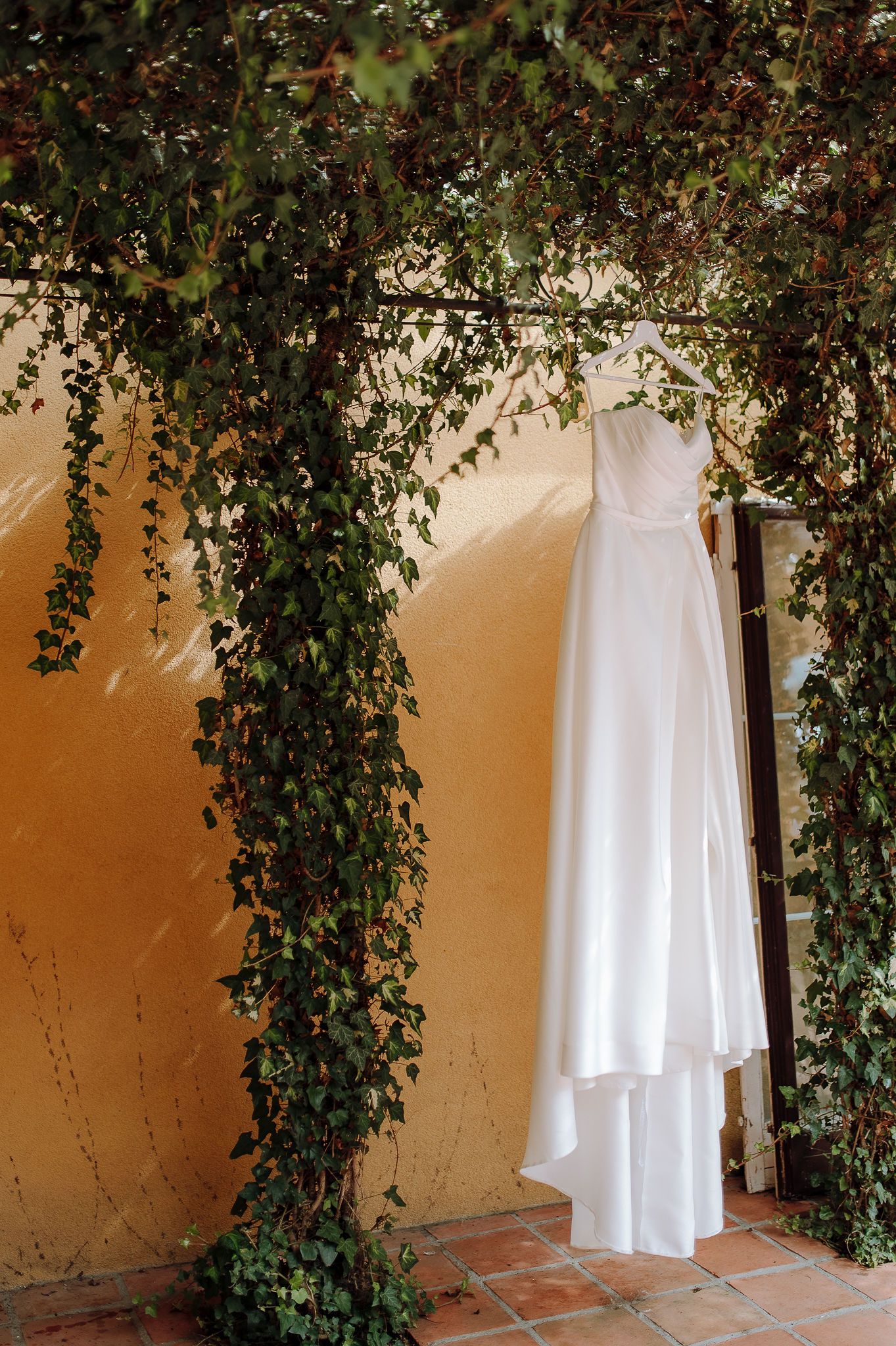 White wedding dress hanging elegantly against an ivy-covered wall with terracotta tiles below.