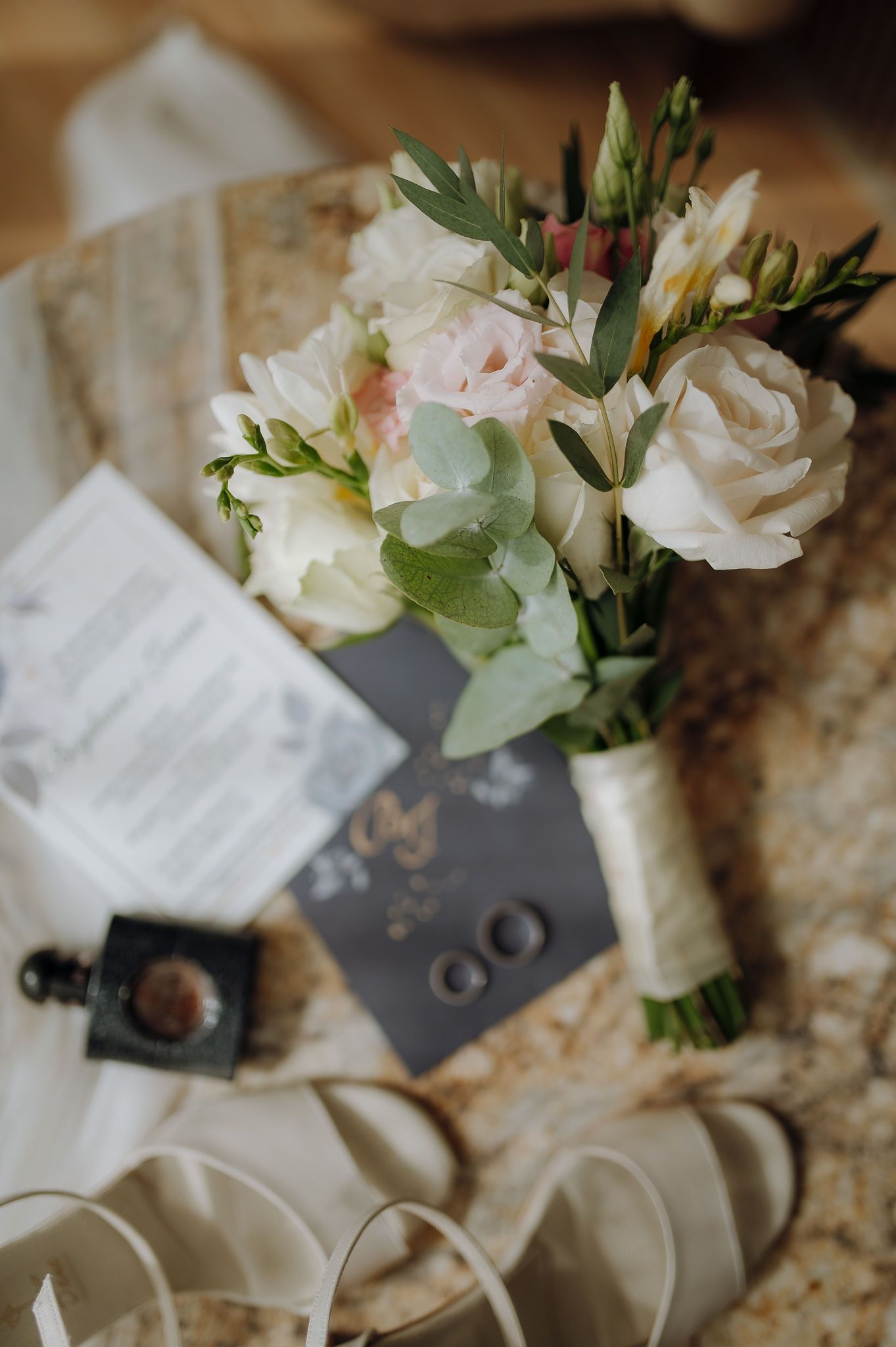 Wedding details with a delicate bouquet of roses and greenery next to rings on a textured surface.