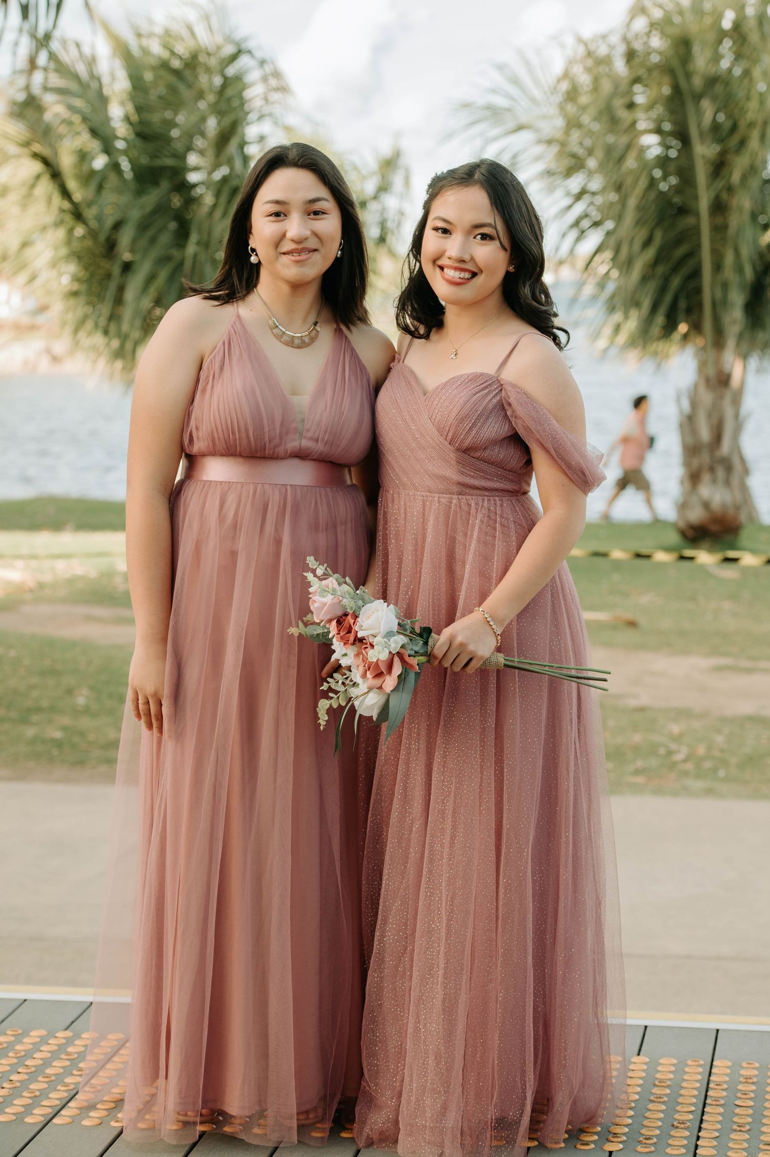 Two bridesmaids in dusty rose tulle dresses stand together holding bouquets at an outdoor wedding venue.
