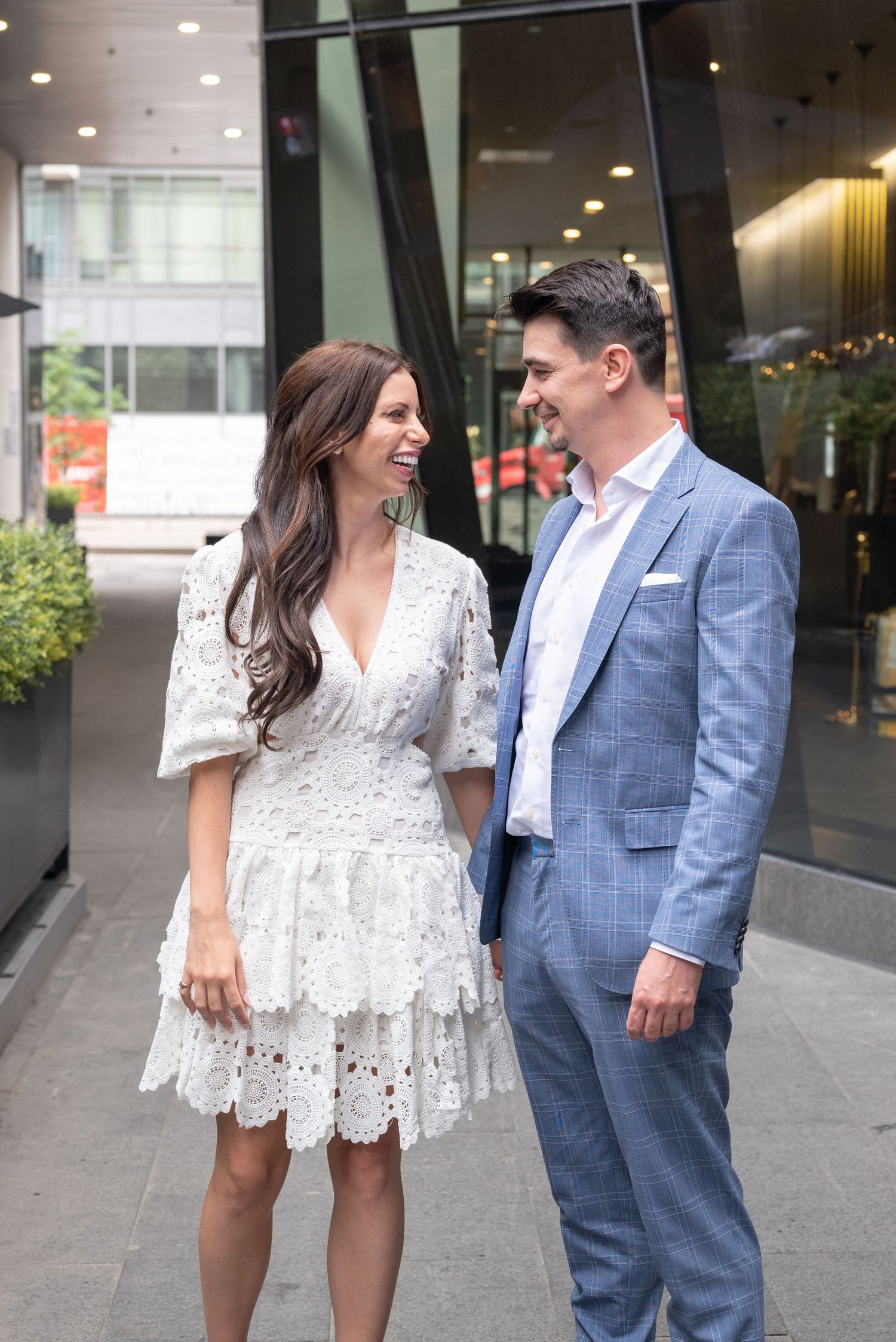 Couple in formal wear share a laugh outside modern building entrance.