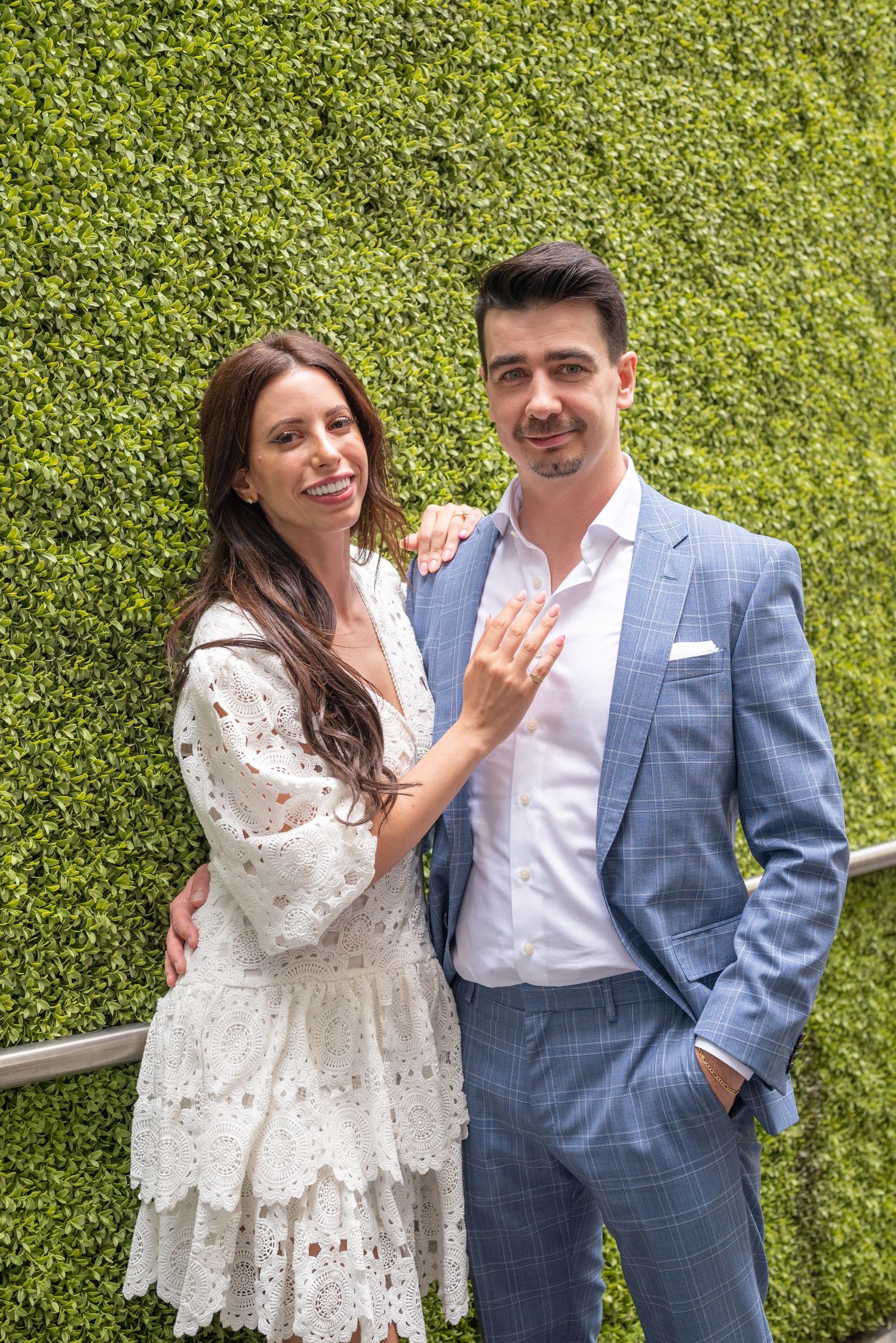 A couple poses in front of a lush green hedge wall showing off an engagement ring.