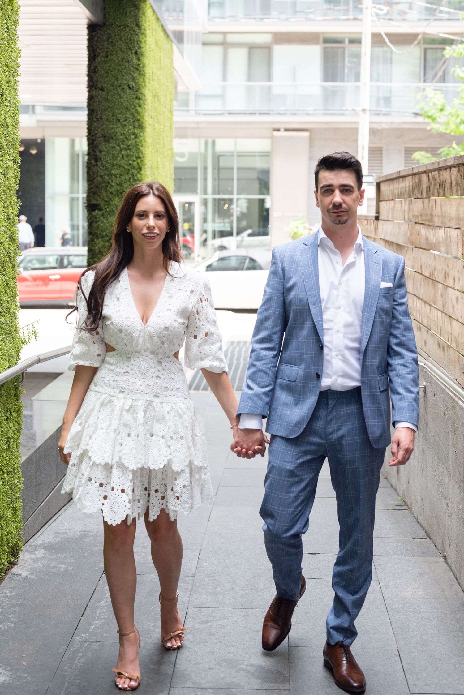 A couple in formal attire walks hand in hand down an urban pathway with green walls.