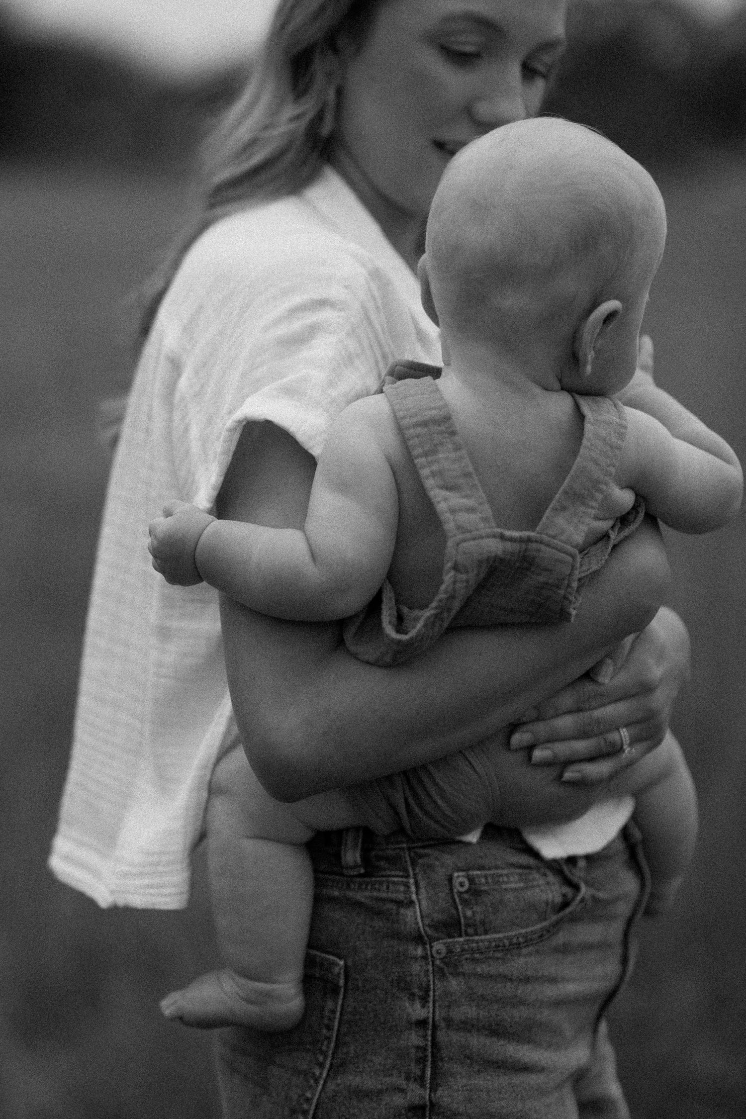 black and white image. back view of a six month old baby as his mom holds him. he is wearing overalls with no shirt