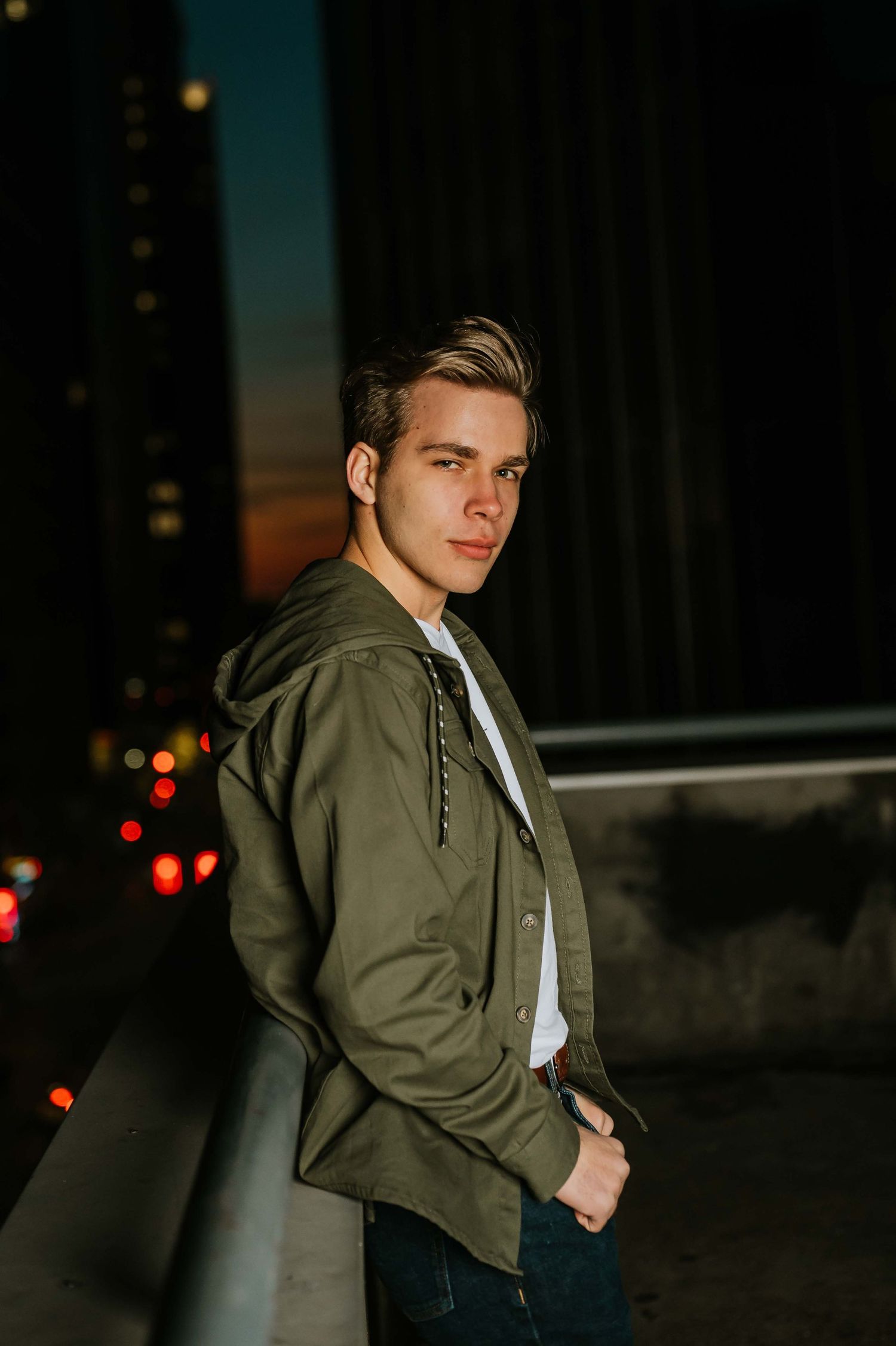 Moody nighttime portrait with person in olive bomber jacket against city lights.