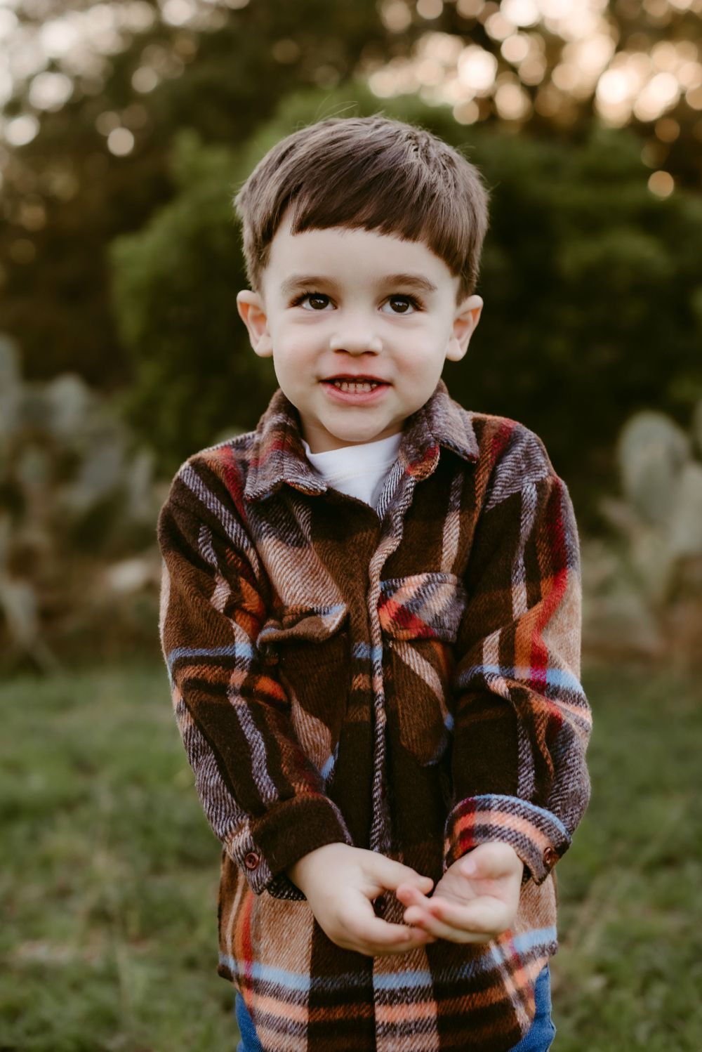 A young child smiles warmly while wearing a plaid jacket in an outdoor setting with bokeh lights.