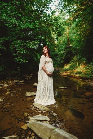 Maternity portrait in white dress standing on rocks by stream in forest setting.