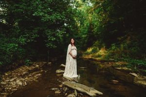 Ethereal maternity photo with woman in white dress by woodland stream.