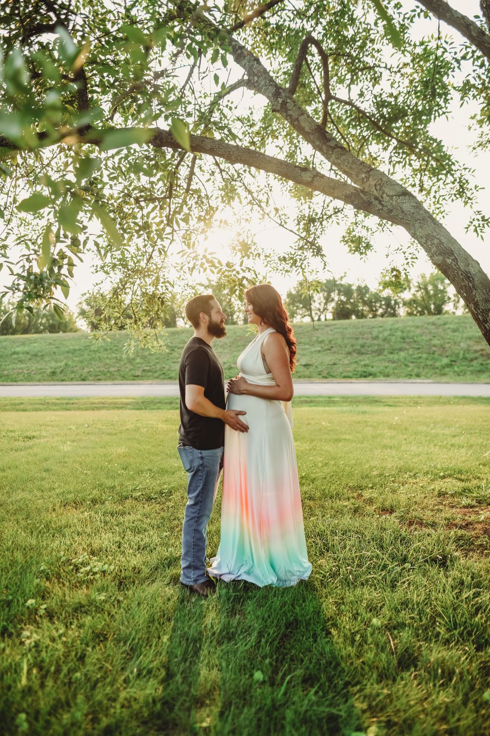 A couple stands under a large tree branch in a sunlit grassy field during sunset.