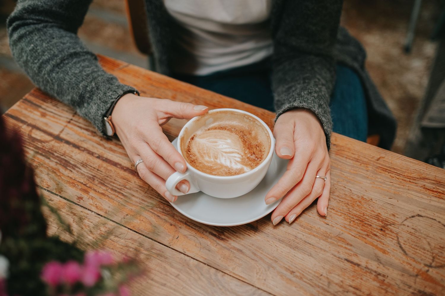 Hands hold a cappuccino with latte art on a wooden table.