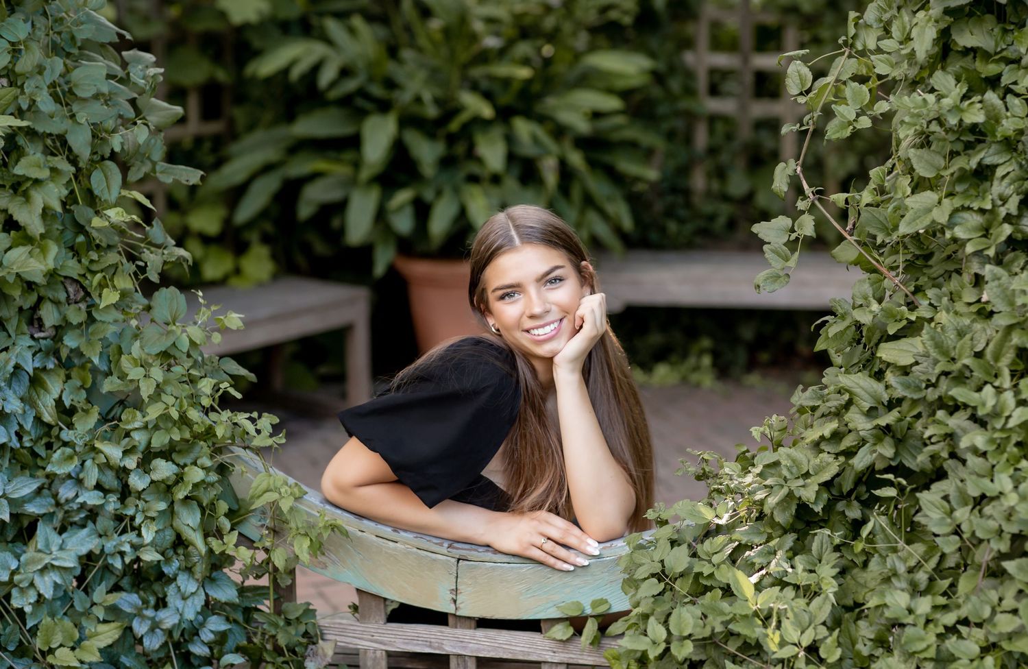 Smiling young woman with long dark hair leaning on a garden chair surrounded by lush green ivy.
