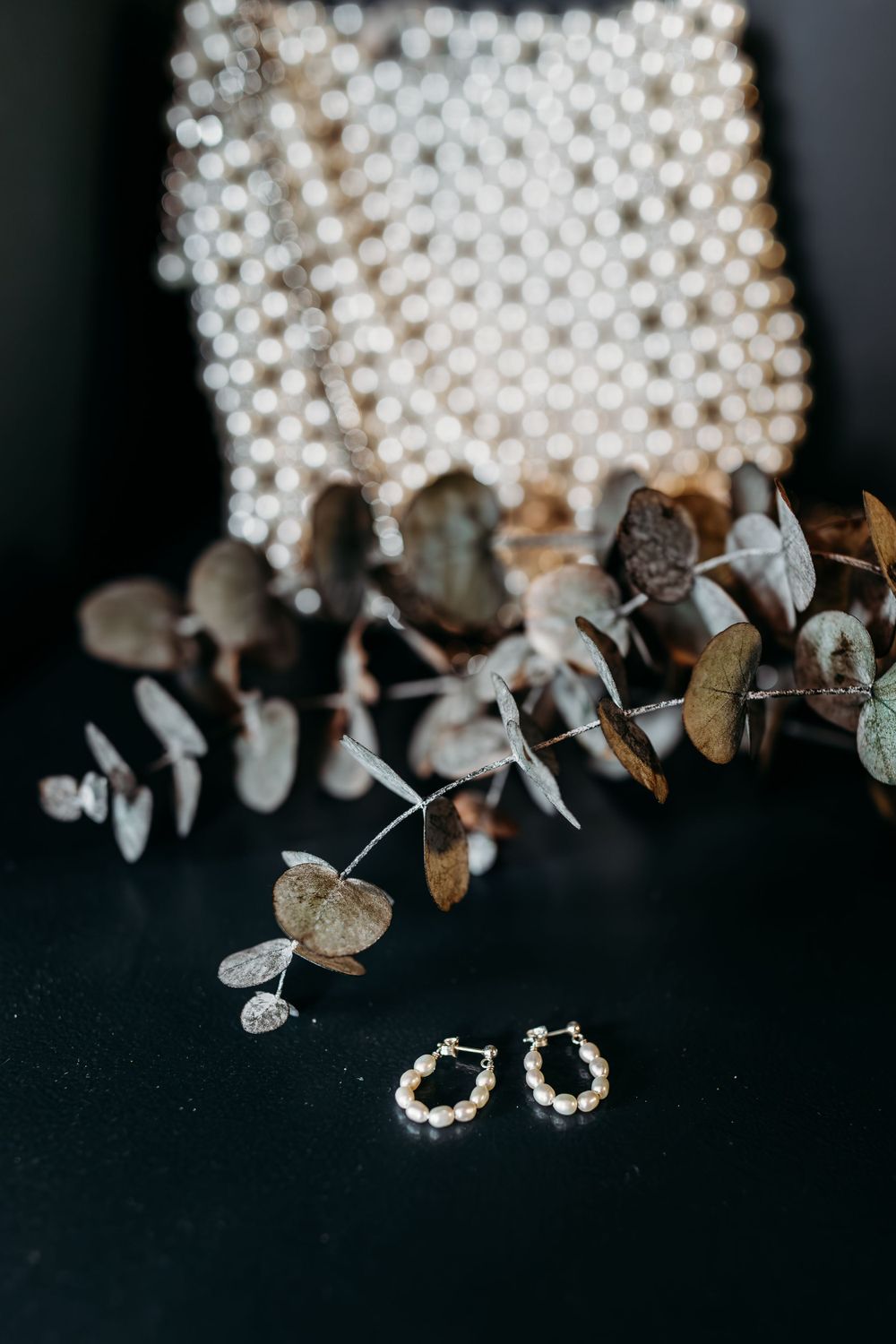 Delicate pearl hoop earrings displayed with dried eucalyptus branches on a black surface.