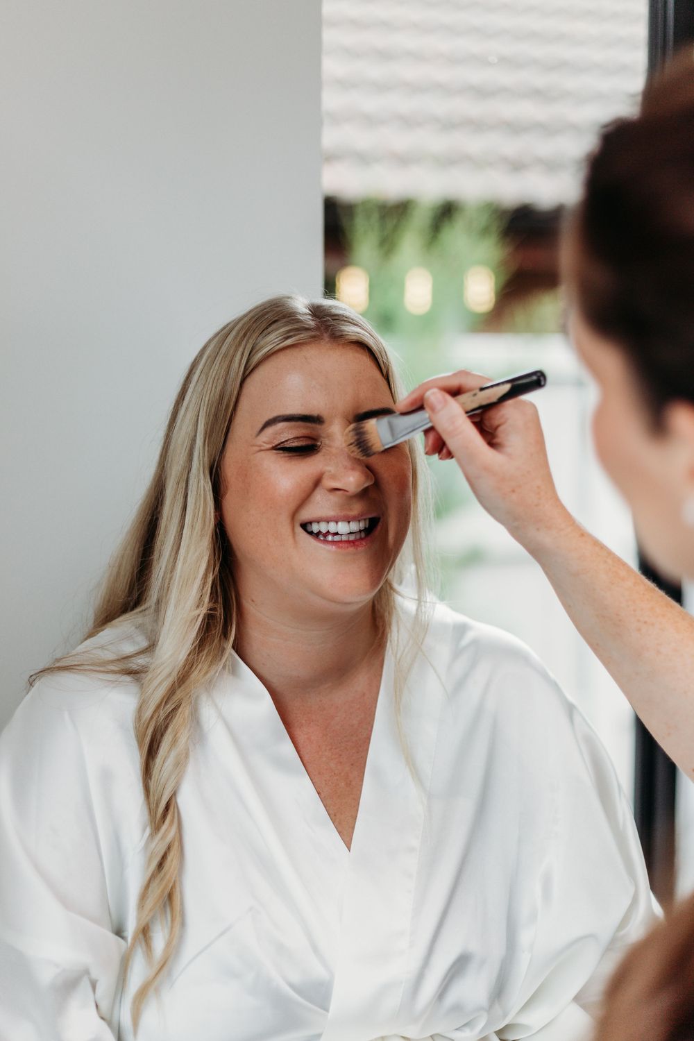 A makeup artist applies eye makeup on a smiling person wearing a white robe during wedding preparations.