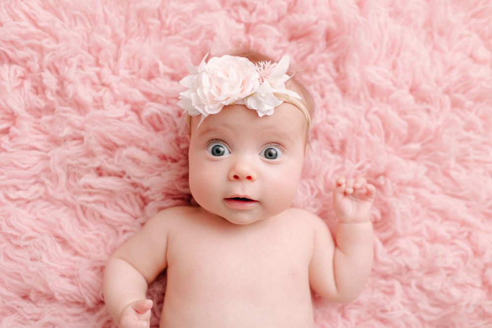 Baby lying on pink furry rug wearing white floral headband against light background.