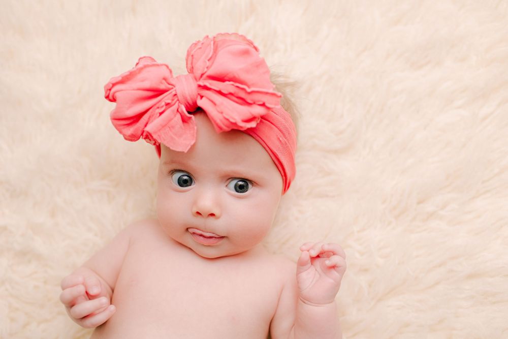 Infant wearing large coral bow headband lying on cream textured backdrop.