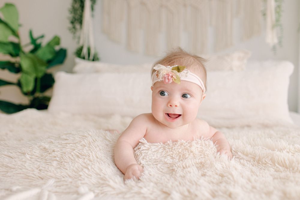 Baby wearing gold headband smiling on white textured bedding with greenery in background.