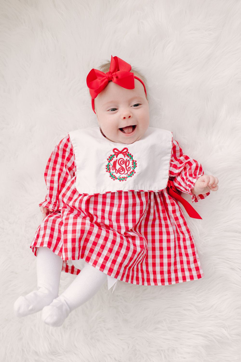 Baby wearing red and white gingham dress with matching bow on plush white background.
