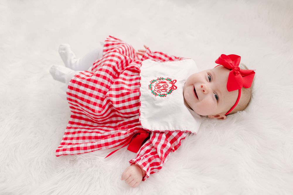 Baby wearing red and white gingham dress with matching bow on white fur background.