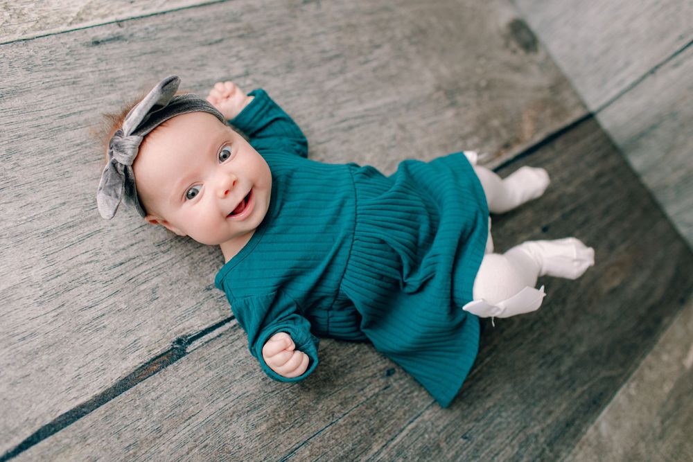Baby wearing teal dress and floral headband lying on wooden surface.