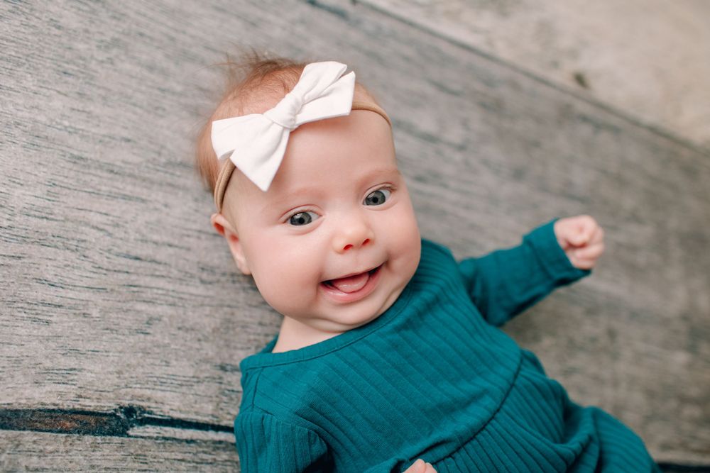 Baby in teal outfit with white bow smiling on wooden floor.